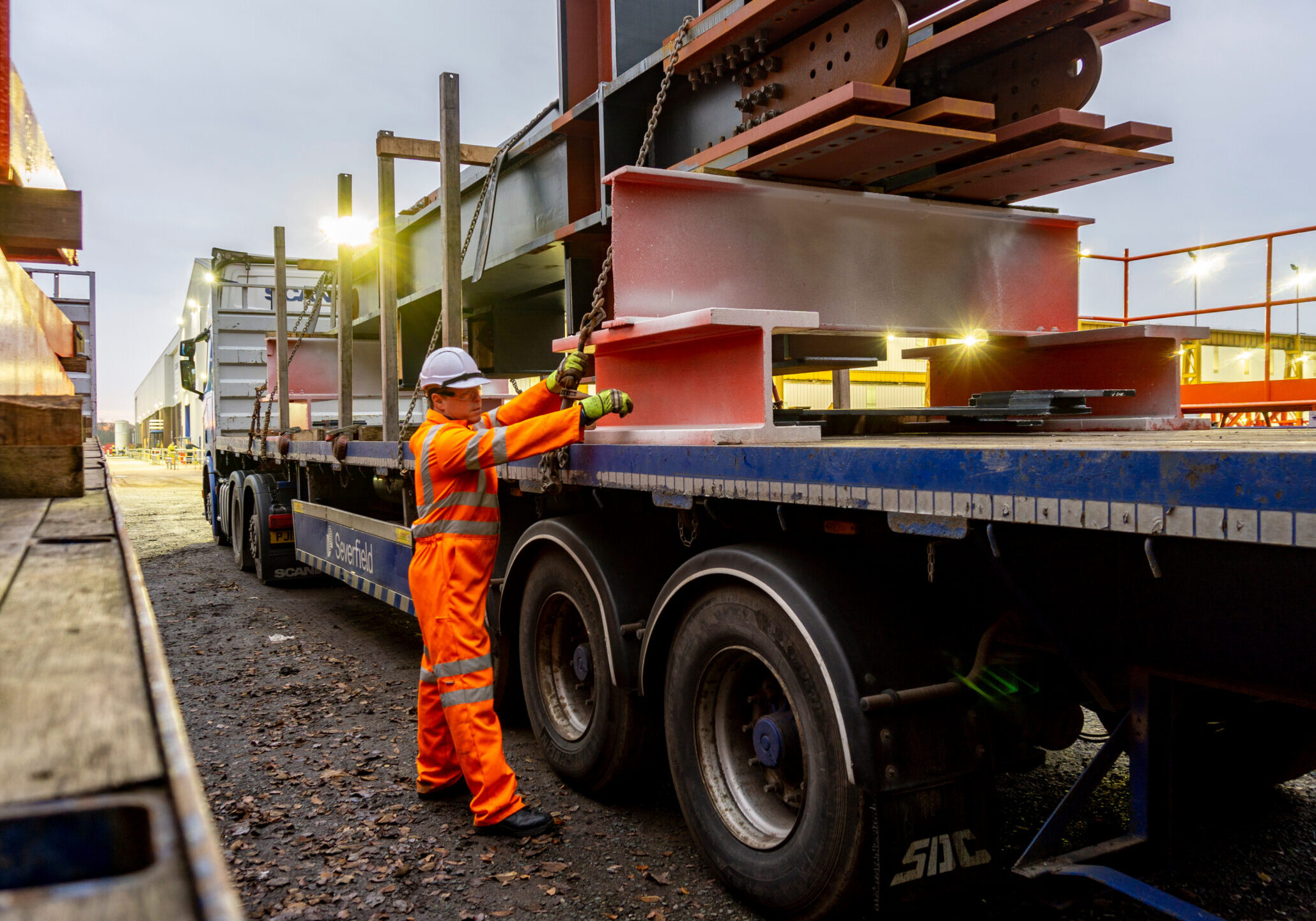 Bolton, Greater Manchester / UK Driver Wearing Full PPE and Chaining Down a Load of Steel Structures onto a Semi Truck Flat bed with Metal Posts Fitted.
